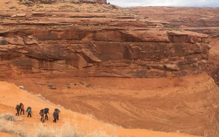 A group of people hike through a desert landscape surrounded by tall canyon walls.